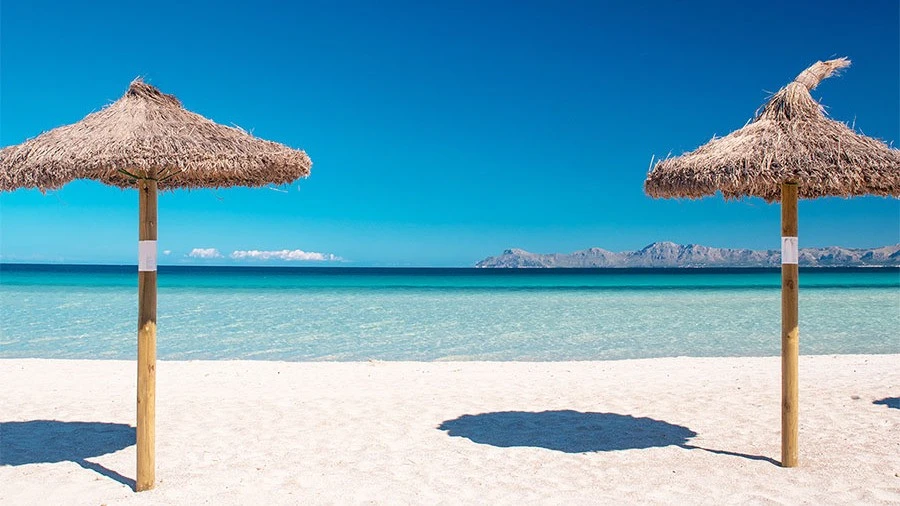 Das Foto zeigt zwei Sonnenschirme aus Stroh am Sandstrand von Mallorca vor dem azurblauen Meer – ein Anblick, der auch die Teilnehmer der Just Dent It Summer Edition erwartet.
