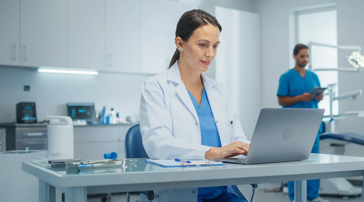 Eine Zahnärztin sitzt in ihrer Praxis am Schreibtisch vor einem Laptop.